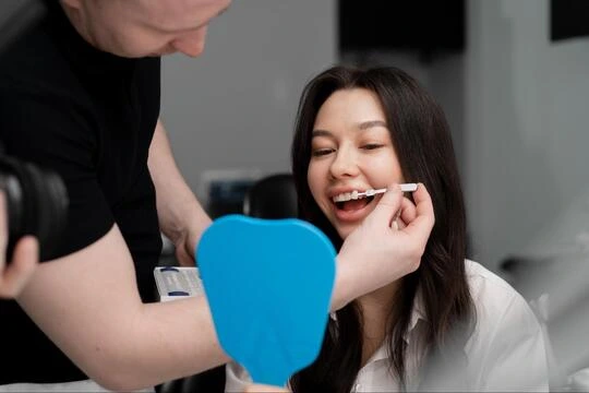 A woman looking into a hand mirror while a dentist checks her teeth after a prosthodontic procedure.
